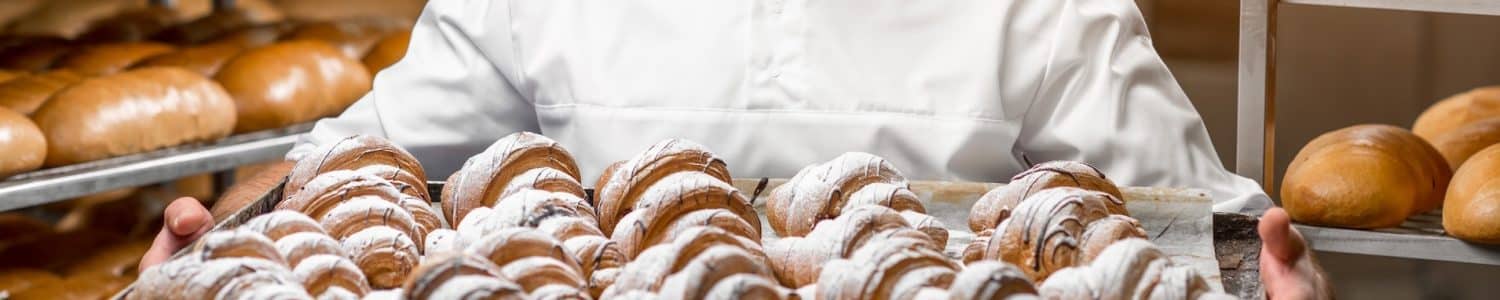 Handsome baker in uniform holding tray full of freshly baked croissants at the manufacturing