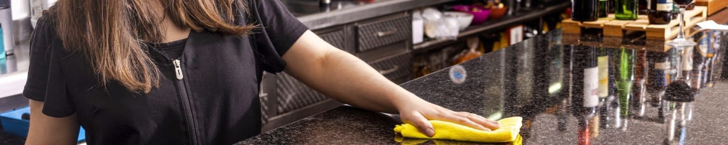Pretty young waitress in apron bending while using detergent and duster to clean it for new guests of restaurant